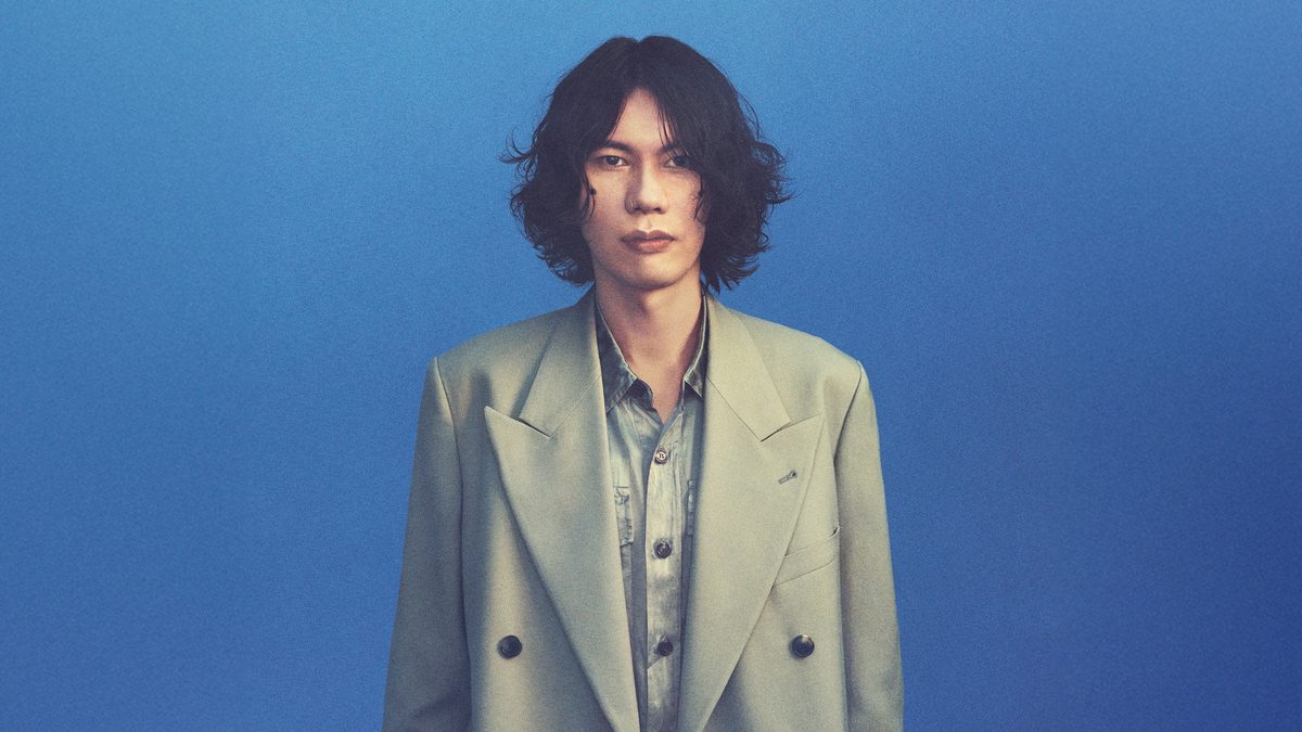 Kenshi Yonezu in a pale jacket against a blue wall, GHOST tour press photo