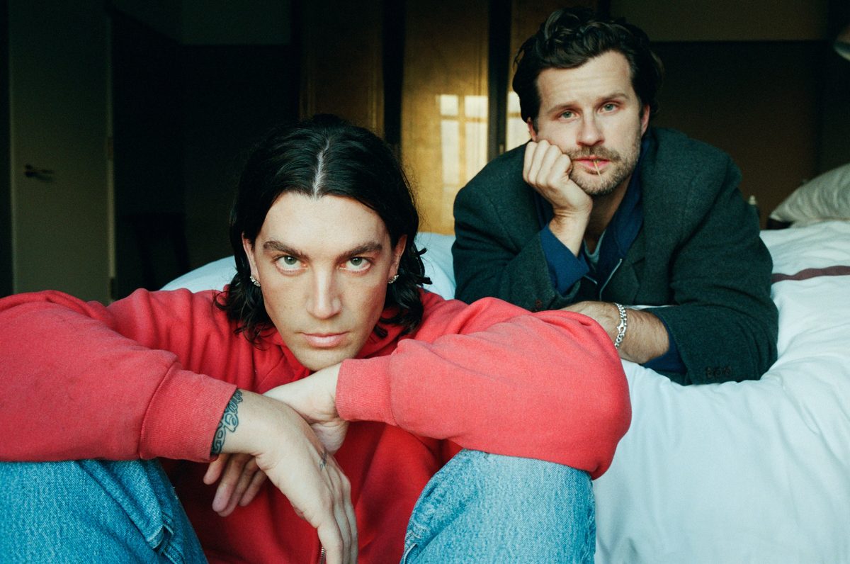 LANY members sitting on a bed, one in a red jumper, soft world tour photo