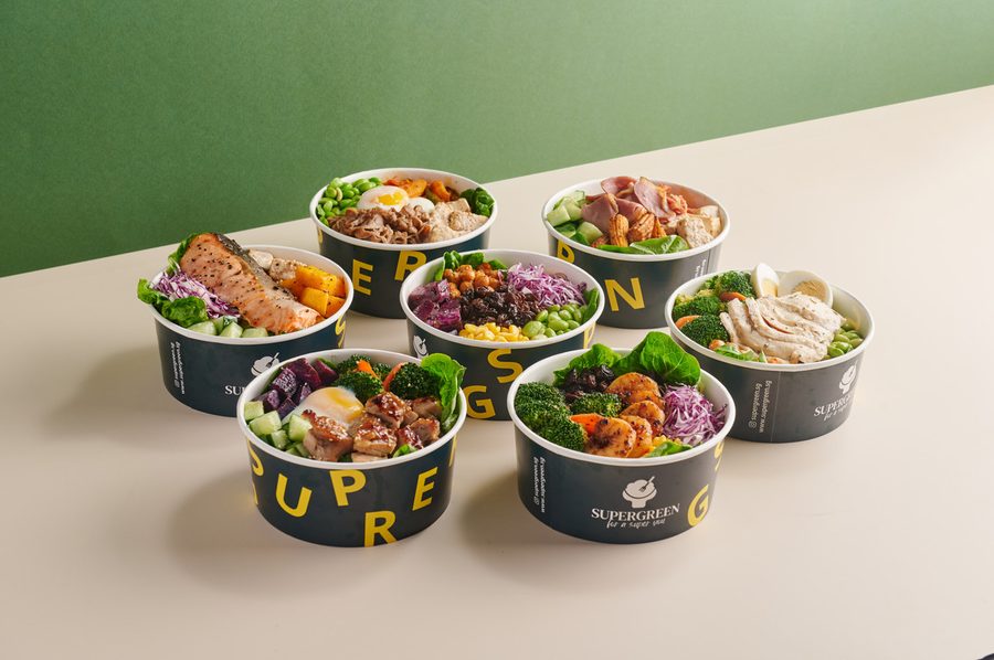 Seven Supergreen salad bowls in the brand's dark green and yellow cups, with salmon, chicken, egg and greens