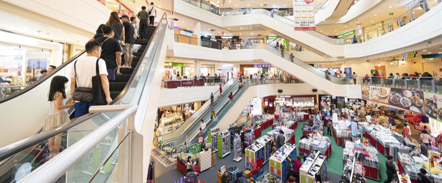Hougang Mall atrium with shoppers on the escalators and a sale fair on the ground floor