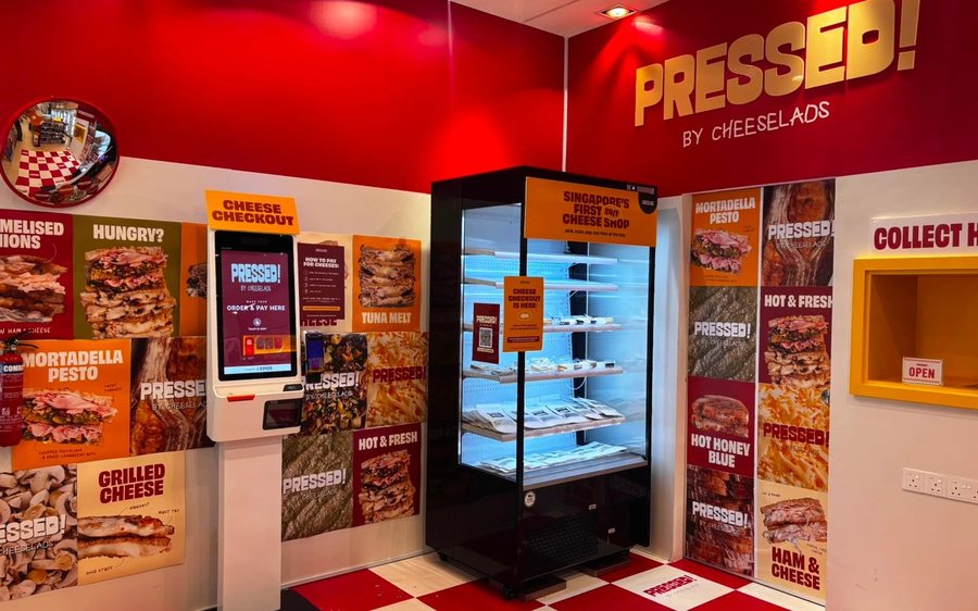 Inside PRESSED! by Cheeselads: gold PRESSED! wall sign, a 24/7 self-checkout cheese fridge, order screen and menu posters