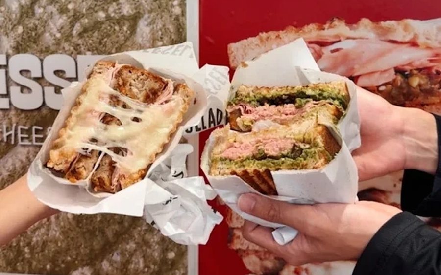 Two PRESSED! grilled cheese sandwiches held up in front of the kiosk menu wall